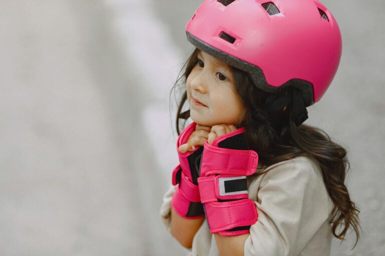 Young girl wearing a bright pink helmet and gloves, providing a side view with a blurred background.