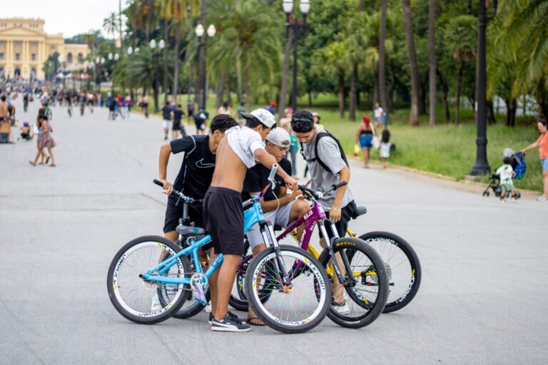 Teenagers gather with bikes in a park, surrounded by trees and lively pedestrians.