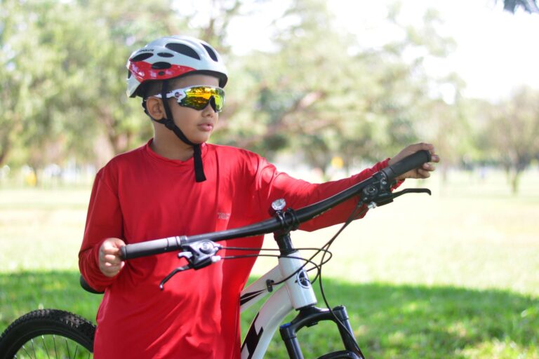 Young boy in a red shirt with a bicycle in a sunny park wearing a helmet and sunglasses.