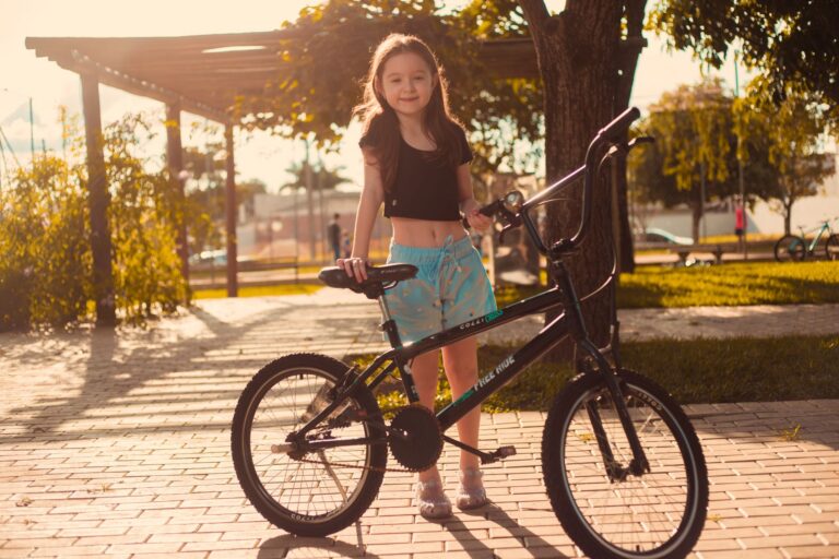 A smiling girl with long hair stands with her bike in a sunny urban park setting.