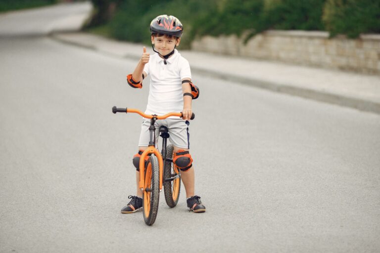 Smiling boy on an orange bike showing thumbs up on a sunny day, wearing safety gear.