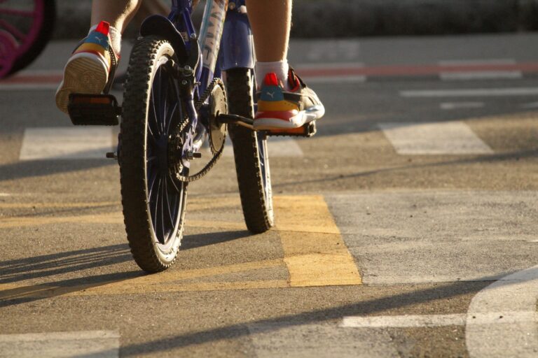 Close-up of child's feet on bicycle pedals in sunlit urban setting.