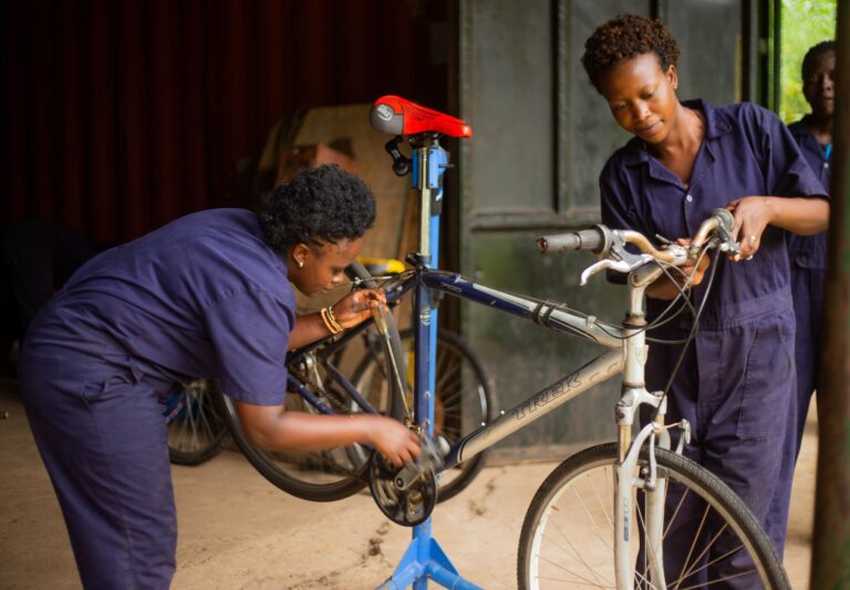 Two female mechanics in blue overalls working on a bicycle inside a workshop.