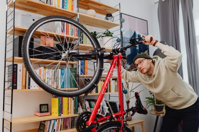 A man in a hoodie adjusts a bicycle indoors in front of a bookshelf.