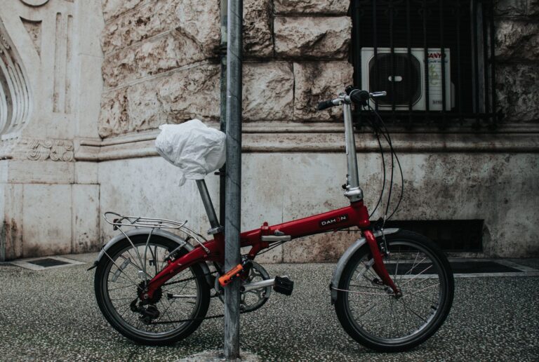 A chained red folding bicycle against a textured stone wall captured on an urban street.
