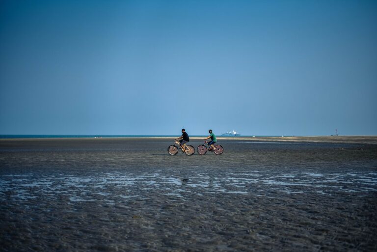 Two men cycling on a beach during low tide under a clear blue sky.