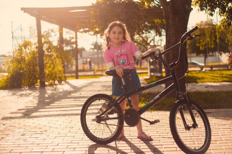 A cheerful girl stands proudly with her bicycle in a sunlit park setting.