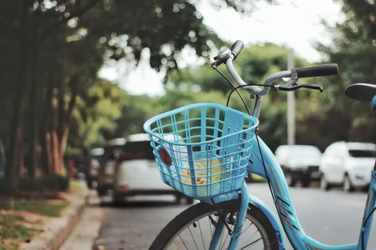 A blue bicycle with a basket parked on a leafy city street during the day.