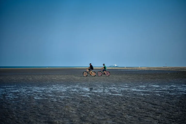 Two men cycling on a beach during low tide under a clear blue sky.